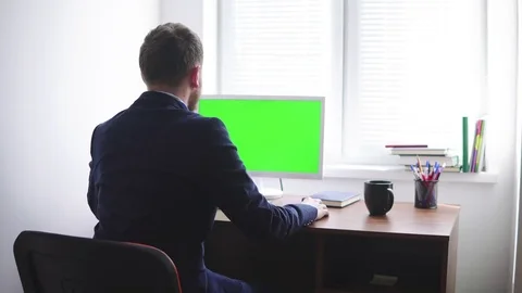 Young man is working on a computer with a mock-up green screen at the table by Stock Footage 75440113