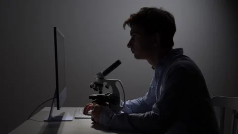 Young man working in a dimly lit laboratory, looking through a microscope while Stock Footage 342673326