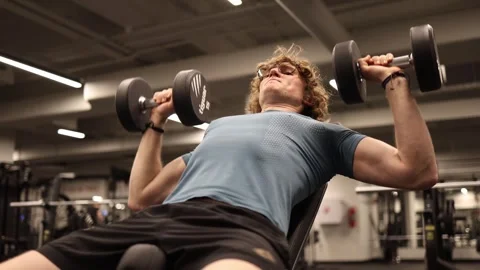 A young man is working on his chest with dumbbells in the gym. He is focused on  Stock Footage 301677188