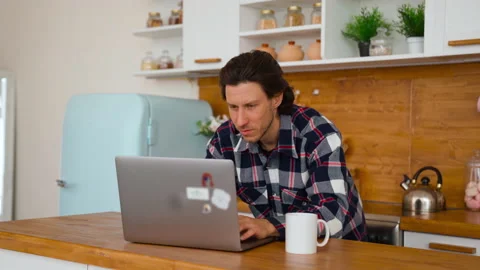 A young man working from home at the notebook in kitchen room. Thinks and typing Stock Footage 137970934