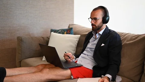 Young man working from home sitting on the sofa with earphones, suit and shorts Stock Footage 128932109