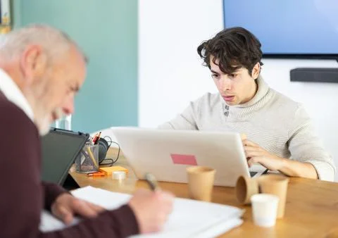 Young man working on laptop, elderly man takes notes on documents Stock-Fotos