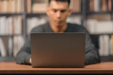 Young Man Working on Laptop in Library with Bookshelf in Background Stockfoto's