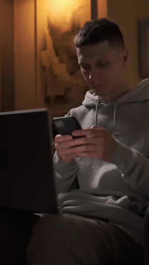 Young man working on laptop in warm evening light, taking smartphone to send Stock Footage 314710272