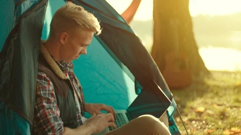 Young man is working at laptop while sitting in the tent at campsite during Stock Footage 98672486