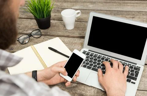 Young man working on laptop while using smartphone at rustic wooden desk in a 스톡 사진