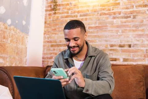 Young man working remotely using laptop and smartphone on sofa Stock Photos