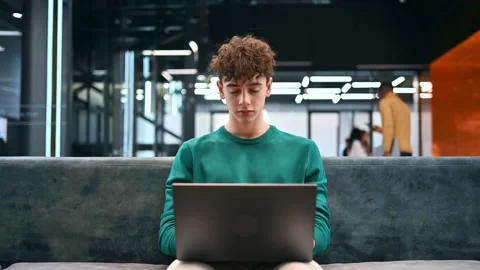 A young man working using a laptop while sitting on a sofa in an office Stock Footage 235232278