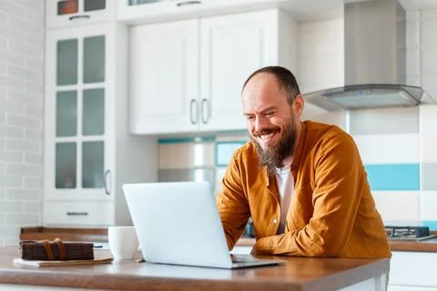Young man working using laptop in kitchen in home interior. Smiling Freelance Stock Photos