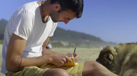 Young man writes in his journal sitting on the beach with his dog Video stock 52257819