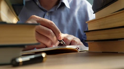 Young man writing in note on a table with piles of books. Knowledge and wisdom. Vidéo 88983541