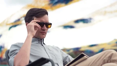 Young man writing a pencil in notebook sitting on chair at city park. Summer day Stock Footage 79356353