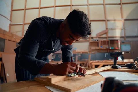 Young man writing with pencil while using tool on wood at woodworking factory Stock Photos