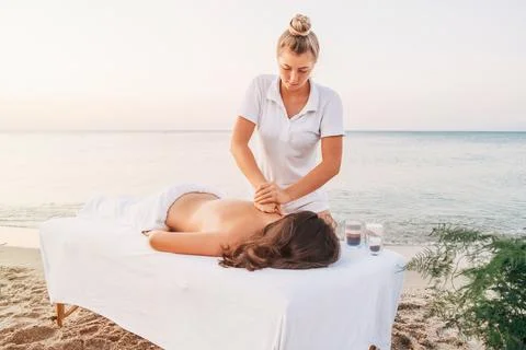 A young masseuse in white clothes makes a massage on the seashore at dawn. Stock Photos