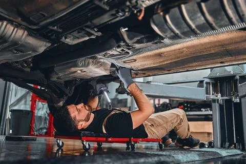 Young mechanic lying down and working under car at the garage. Stock Photos