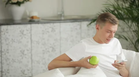 Young men sitting on sofa eating apple and using cellphone Видео 27691917