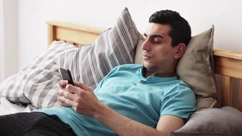Young middle eastern male using a smartphone, lying on bed at home Stock Footage 86402415