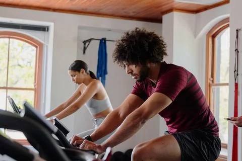 Young middle eastern man and biracial woman cycling on exercise bike at the gym Stock Photos