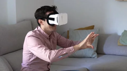 Young millennial man in virtual reality headset sitting on couch at home work Stock Footage 145775640
