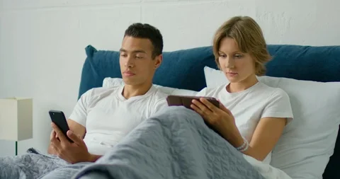 Young mixed couple lying in bed in the morning, using gadgets Stock Footage 113642117