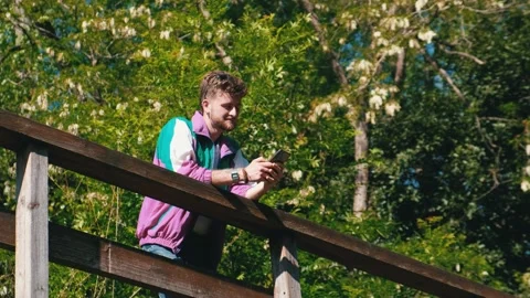 Young modern guy talking on the phone and sending a message on a wooden bridg Stock Footage 192898119