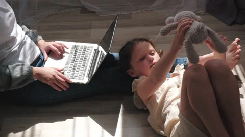 Young mom works at computer near little daughter playing with fluffy bear Stock Footage 200913751