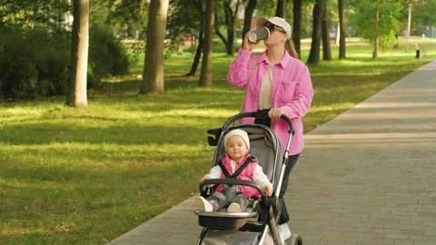 Young mother drinks coffee while walking with her child in an outdoor park, a Stock Footage 234317507