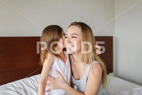 Young mother hugging little daughter, sitting on bed with white linens. Stock Photos