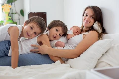 Young mother lying in bed with her newborn baby boy and his older brothers Stock Photos