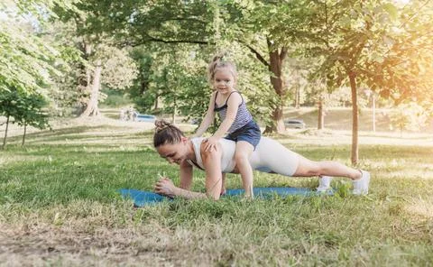 A young mother performs a plank exercise with her little daughter on her back Stock Photos