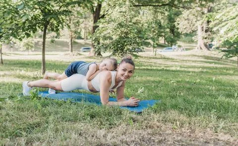 A young mother performs a plank exercise with her little daughter on her back Stock Photos