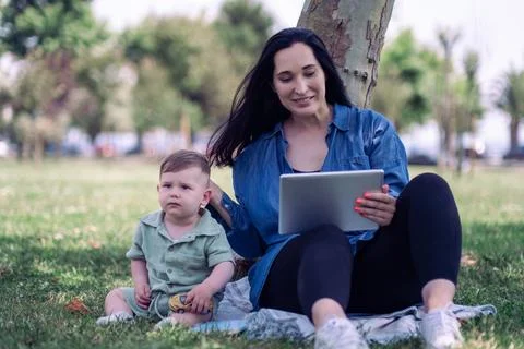 Young mother with tablet computer sitting with little son on lawn grass in Stock Photos