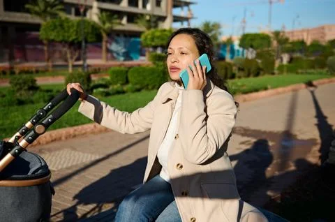 Young mother talking on cellphone while pushing stroller in park on a sunny d Stock Photos