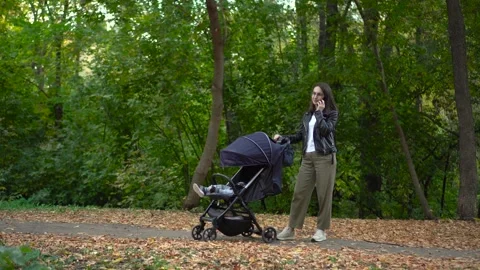 A young mother is talking on the phone and rocking her baby in a stroller. A Vídeos de archivo 252129287