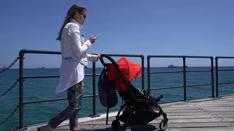 Young mother types a message on cell phone while walking along a sea quay with Video stock 76989755
