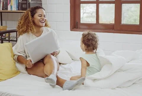 Young mother using laptop for work near playing daughter on bed. Foto stock