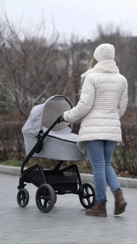 Young mother in white puffer jacket and beanie pushes modern baby stroller  Video stock 341834913