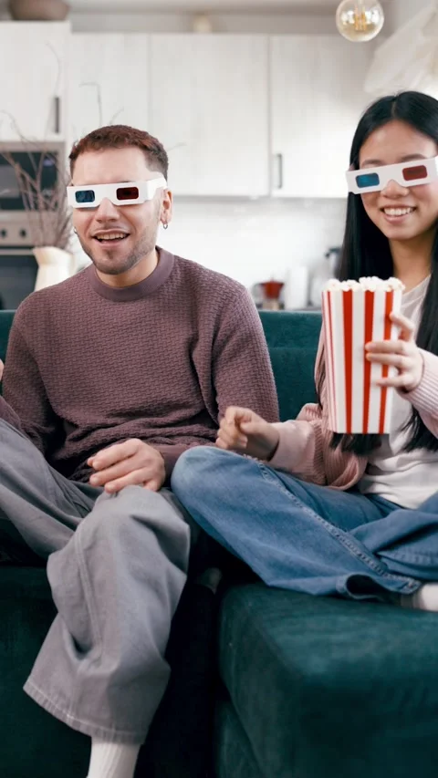 Young multicultural couple watching a 3d movie at home eating popcorn. Vertical Vídeos de archivo 339032711