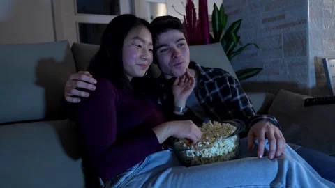 Young multiethnic couple sharing popcorn on sofa at night Stock Footage 305236998