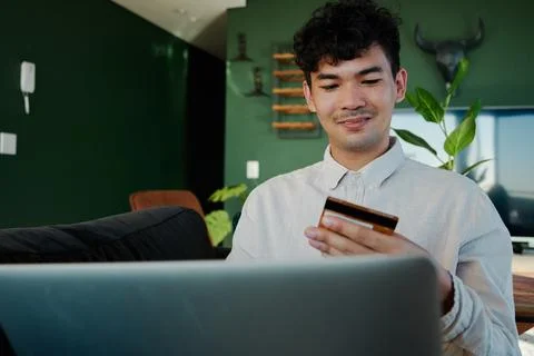 Young multiracial man in shirt using laptop and credit card in living room at Stock Photos