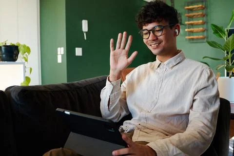 Young multiracial man smiling while using digital tablet and waving on sofa at Stock Photos
