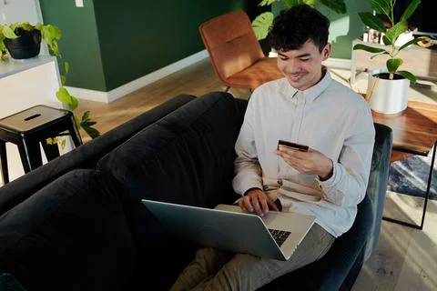 Young multiracial man smiling while using laptop and credit card on sofa at home Stock Photos