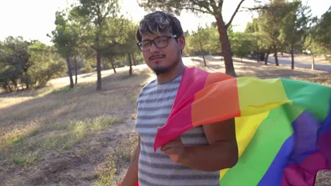 Young non-binary guy wrapped in LGBTQ flag suddenly looking at camera 스톡 동영상 250466355