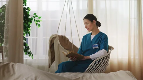 Young nurse reading a book while sitting near a sleeping patient in care home Stock Footage 327987107