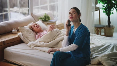 Young nurse talking on cellphone while neglecting senior patient in bed Stock Footage 327987199