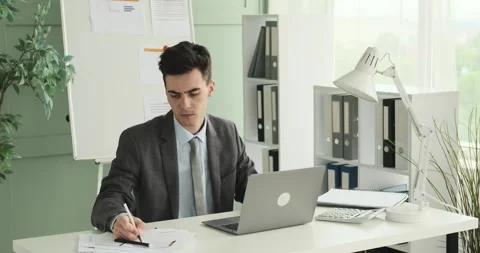 Young Office Worker Handling Papers and Typing on Laptop in Office Stock Footage 248533608