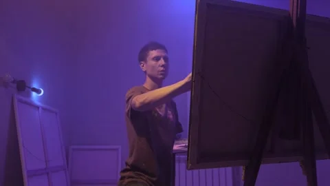 Young painter creates art in studio under purple atmospheric light Stock Footage 312330067