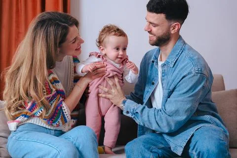 Young parents smiling while helping baby girl stand on sofa during family time Stock Photos