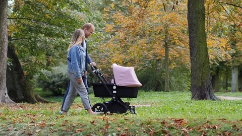 Young parents walk roll a stroller with baby child on a city park alley Stock Footage