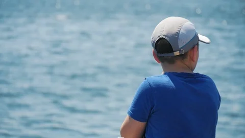 Young pensive boy in white cap and blue t-shirt looking on the sea's water with Video stock 111157066